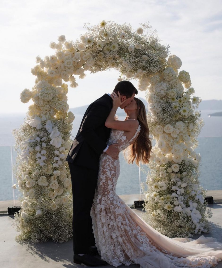 Newlywed couple embracing in front of white floral arch.