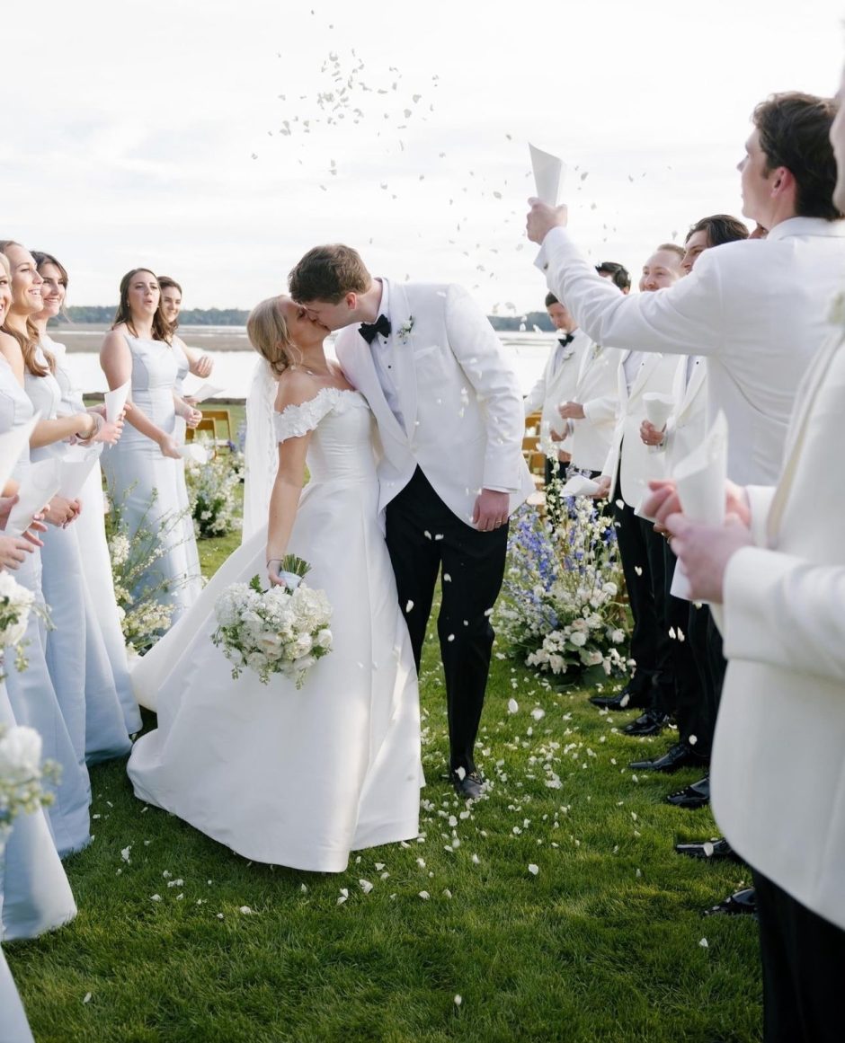 A newlywed couple sharing a kiss at their outdoor wedding ceremony, surrounded by their bridal party dressed in soft blue and white attire. Guests are showering them with flower petals as they celebrate near a serene lake backdrop.