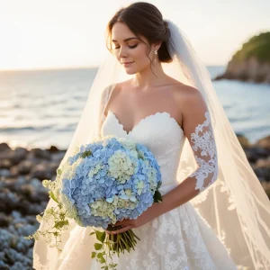Bride standing on the beach holding a bouquet of light blue hydrangeas, showcasing dreamy pastel wedding flowers perfect for spring and summer weddings.