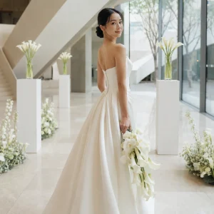 East Asian bride in modern ivory gown holding white calla lily bouquet in a bright glass-and-stone wedding venue.