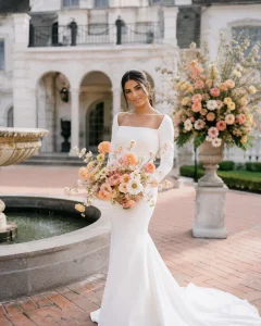 Iberian bride with warm-toned skin holding a Butterfly Ranunculus bouquet in blush, peach, cream, apricot, coral, and buttery yellow at an Ashford Estate-inspired wedding, showcasing airy branching blooms and elegant garden-style floral design.
