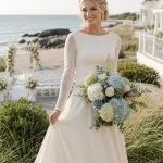 Smiling Alpine bride with braided honey-blonde hair holding a white, pale blue, and soft green hydrangea wedding bouquet at an Inn by the Sea-inspired coastal venue.